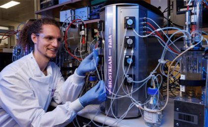 A man in a white coat and blue gloves sits in front of a silver cannister 